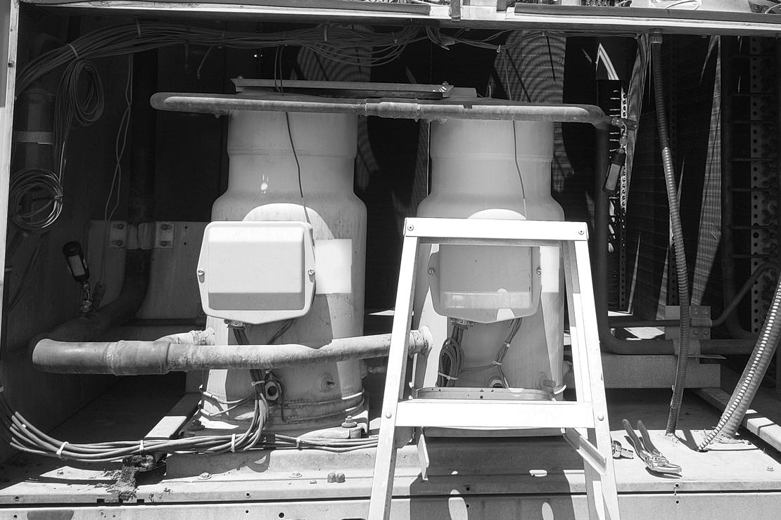 Two scroll compressors in the opened compressor compartment of a rooftop unit, a stepladder in front of them and the pipework around them
