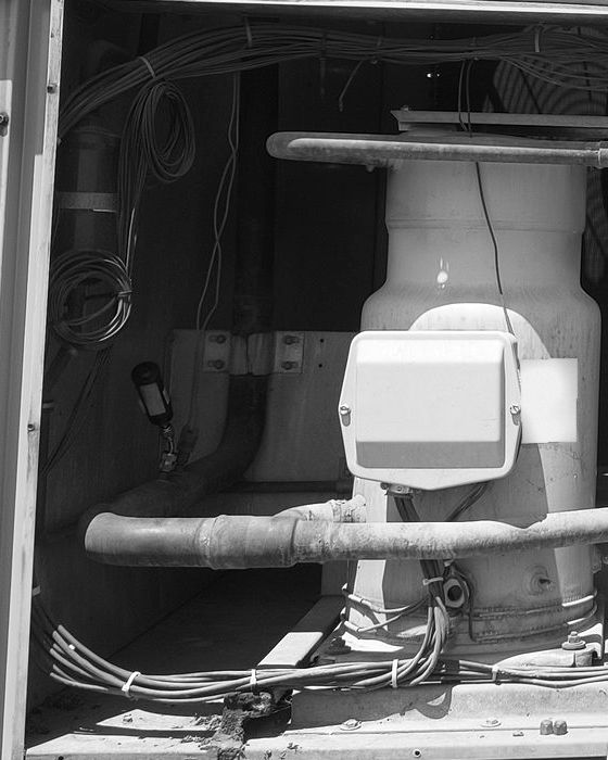 A scroll compressor standing in the opened compressor compartment of a rooftop unit, its terminal box facing out, with a pipe and a bundle of wiring in front of it
